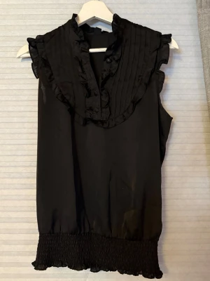 Svart ärmlös blus med volang - Stilren svart blus med volangdetaljer och knappar framtill. Blusen är ärmlös och har en smockad nederkant som ger en snygg siluett. Materialet känns lätt och svalt, perfekt för en trendig look. Passar till både jeans och kjol.