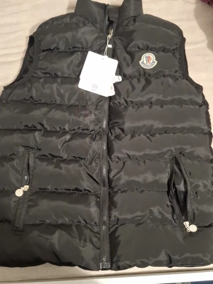 Svart dunväst från Moncler - Snygg svart dunväst från Moncler med klassisk logga på bröstet. Västen har två dragkedjeförsedda fickor och quiltad design. Perfekt för lager-på-lager och håller dig varm med lätt dunfyllning. Stängs med dragkedja framtill.