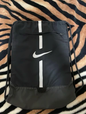 Svart gympåse från Nike - Snygg svart gympåse från Nike med vitt logotryck och vertikal rand på framsidan. Väskan har ett dragkedjefack framtill och snörstängning som även fungerar som axelband. Tillverkad i slitstarkt syntetmaterial, perfekt för träning eller skola.