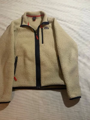 Beige teddyjacka från Patagonia - Säljer en beige teddyjacka från Patagonia i storlek XXL. Jackan har hel dragkedja framtill, två sidofickor med dragkedja och detaljer i mörkblått samt röda dragkedjor. Mjuk och fluffig med hög krage för extra värme. Perfekt nu för våren.