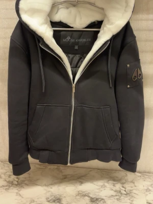 Moose Knuckles bunny S - Svart teddyjacka från Moose Knuckles med vit fluffig fuskpäls på insidan och huva. Jackan har dragkedja framtill, två stora fickor och en patch med logga på ärmen. Perfekt för kyliga dagar och riktigt snygg streetstil.