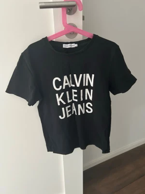 Svart Calvin Klein Jeans t-shirt - Snygg svart t-shirt från Calvin Klein Jeans med vit logotyp framtill. Klassisk rund halsringning och korta ärmar. Tillverkad i mjuk bomull som känns skön mot huden. Perfekt för en avslappnad och trendig look.