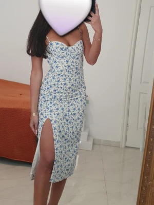 Blommig vit och blå klänning från Bershka - Säljer en söt vit klänning med blått blommönster och smala axelband. Klänningen har en figurnära passform, markerad byst och en hög slits på ena sidan. Perfekt för sommarens alla tillfällen!