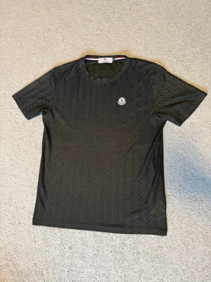 Svart mönstrad t-shirt från Moncler - Säljer en svart t-shirt från Moncler med diskret zigzag-mönster och broderad logotyp på bröstet. Den är in ett unnar matrial perfekt som träningströja. Väldigt bra skick då jag knappt har använt den