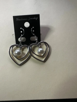 Silvriga hjärtörhängen med pärlor - Säljer ett par snygga örhängen med dubbla hjärtan i silverfärgad metall och vita pärlor som hänger i mitten. Perfekt accessoar för dig som gillar romantisk och trendig stil. Krokar i metall för enkel användning.