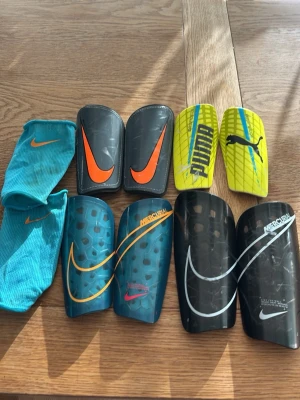 Benskydd Nike och Puma, flera färger - Säljer flera par benskydd från Nike och Puma i olika färger: svart, blå, gul och turkos. Några har Mercurial-design och stora loggor. Ett par turkosa strumpor från Nike ingår. Perfekta för fotbollsträning eller match.