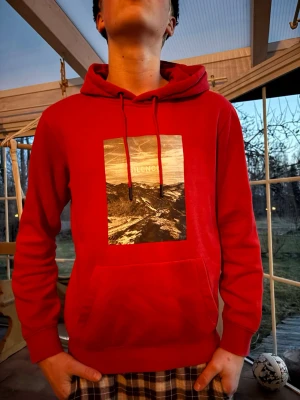 Röd hoodie med tryck 'Silence' - Röd hoodie med stor svartvit fotoprint på bröstet där det står 'SILENCE'. Hoodien har huva med dragsko och en stor magficka. Perfekt för dig som gillar streetstyle och vill sticka ut med en cool print. Mjuk och skön bomullskänsla.