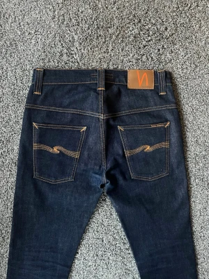 Mörkblå jeans från Nudie Jeans - Snygga mörkblå jeans från Nudie Jeans med klassisk femficksdesign och kontrastsömmar. Jeansen har smal passform och ikoniska broderade bakfickor samt läderpatch med logga i midjan. Perfekta för en stilren och modern look.