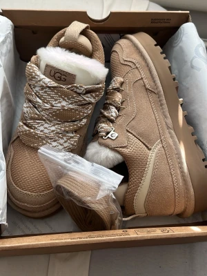 Beige sneakers från UGG med pälsfoder - Orginal pris 1950kr. Snygga beige sneakers från UGG med grov sula och pälsfoder. Skorna har stickade och mockadetaljer, samt mönstrade snören i vitt och beige. Perfekta för dig som vill ha både stil och komfort under kyligare dagar.