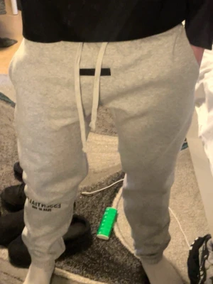 Grå mjukisbyxor från Essentials - Snygga grå mjukisbyxor från Essentials med dragsko i midjan och tryck på ena benet. Byxorna har en avslappnad passform och muddar vid bensluten. Perfekta för en chill och trendig look.