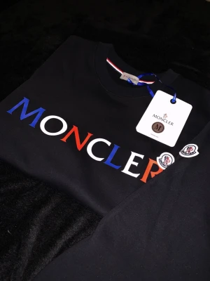 Svart Moncler t-shirt med logga - Snygg svart t-shirt från Moncler med stor logga i blått, vitt och rött över bröstet. Klassisk rund halsringning och korta ärmar. Perfekt för dig som gillar exklusiva streetwear-plagg och vill sticka ut med en ikonisk design.