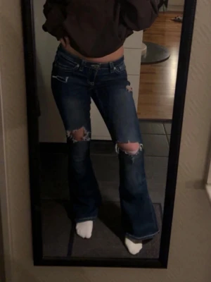 Mörkblå bootcut jeans med slitningar - Säljer ett par mörkblå bootcut jeans med stora slitningar över knäna och lätt slitna detaljer vid fickorna. Jeansen har låg midja och klassisk femficksdesign. Har en mindre slitning nertill som kan visas vid önskan