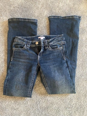 Blå bootcut jeans med broderade fickor - Säljer ett par blå bootcut jeans med vita kontrastsömmar och snygga broderade bakfickor i vitt och beige. Jeansen har klassisk femficksmodell och normal midja. Perfekta för dig som gillar detaljer och en avslappnad men trendig stil.