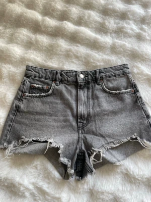 Grå denimshorts med fransig kant - Snygga grå jeansshorts i denim med rå, fransig kant och klassisk femficksdesign. Shortsen har hög midja och en avslappnad passform. Perfekta för sommardagar och festivaler.