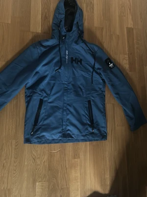 Blå Helly Hansen vindjacka med huva - Snygg blå vindjacka från Helly Hansen med svart HH-logga på bröstet. Jackan har huva med dragsko, två svarta dragkedjefickor och justerbara snören. Perfekt för blåsiga dagar och streetwear-stil.