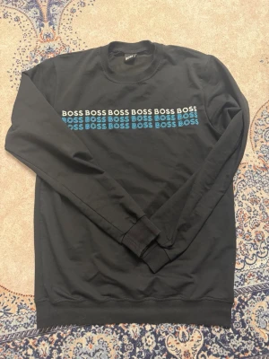 Svart sweatshirt från BOSS - Cool svart sweatshirt från BOSS med tryck i vitt och turkost över bröstet där det står 'BOSS' flera gånger. Tröjan har rund halsringning och långa ärmar med ribbade muddar. Perfekt för en avslappnad och trendig look.