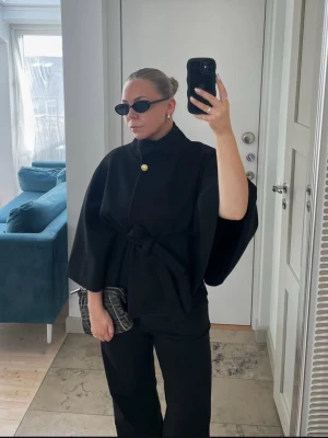 Svart cape-kappa med knytband - Stilren svart cape-kappa med knytband i midjan och en dekorativ guldknapp vid halsen. Kappan har vida ärmar och hög krage, vilket ger en elegant och modern look. Perfekt för dig som vill ha något trendigt och unikt i garderoben.