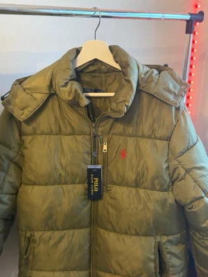 Olivgrön pufferjacka från Polo Ralph Lauren - Snygg olivgrön pufferjacka från Polo Ralph Lauren med dragkedja, huva och två sidofickor. Jackan har det klassiska röda Polo-logot på bröstet och breda quiltade paneler. Perfekt för kalla dagar och riktigt trendig look.