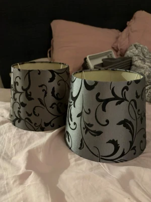 Två grå lampskärmar med svart mönster - Säljer två snygga lampskärmar i grått tyg med svart, blommigt och snirkligt mönster. Skärmarna har en klassisk, lätt konisk form och passar perfekt till mindre lampfötter. Ger ett stilrent och elegant intryck i rummet.