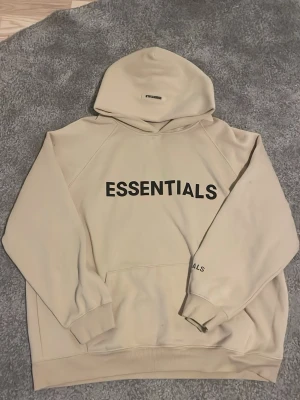 Beige Essentials hoodie med tryck - Snygg beige hoodie från Essentials med stor svart logga på bröstet och mindre tryck på huvan. Klassisk känguruficka framtill och ribbade muddar vid ärmar och nederkant. Perfekt oversized fit och mjukt material för en chill look. Priset kan diskuteras.