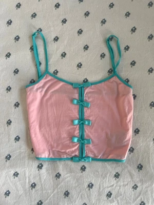 Hatsune Miku Rosa topp med rosetter - Supersöt hatsune miku x dolls kill limited edition croppad topp i ljusrosa med tunna axelband och turkosa kanter. Framtill sitter fyra små rosetter i samma turkosa färg som ger en gullig och unik look. Äxelbandar är justerbara. Materialet känns mjukt och stretchigt, perfekt för sommarens outfits. Storlek M. Måtten finns i bilderna. 
