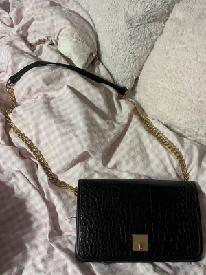 Svart axelväska med kedja - Stilren svart axelväska med croco-mönster och guldfärgad kedja. Väskan har ett elegant metallspänne framtill och en kombinerad axelrem i kedja och läder. Perfekt för dig som gillar trendiga accessoarer.