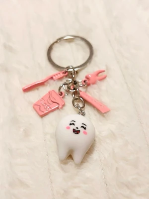 Nyckelring med tand och tandvårdshängen - Söt nyckelring med en vit tand med glad ansikte, samt rosa hängen i form av tandborste, tandtråd och tandpetare. Perfekt accessoar för dig som gillar unika detaljer och tandvård. Metall och plast i rosa och vitt.