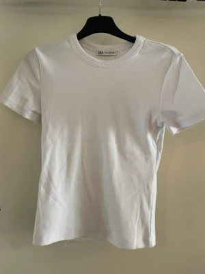 Vit ribbad tight T-shirt från Zara - Klassisk vit t-shirt från Zarai storlek M men passar även som S.  Den har ribbad struktur och rund hals. T-shirten har korta ärmar och är tillverkad i mjuk bomull för en skön känsla. Enkel och stilren modell som passar till allt. T-shirten är ny och har kvar prislappen