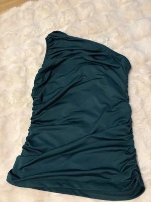 Mörkgrön one shoulder topp Bubbleroom - Snygg mörkgrön oneshoulder topp från Bubbleroom. Toppen har en tight passform och är tillverkad i ett mjukt, stretchigt syntetmaterial. Perfekt för dig som vill sticka ut med en cool och modern look. Toppen är stretchig och passar även S