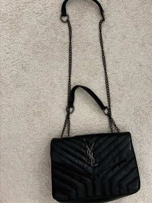 Svart axelväska från Saint Laurent (inte äkta) - Snygg svart axelväska i ”märket” Saint Laurent med quiltat mönster och silverfärgat YSL-logo på framsidan. Väskan har kedjerem med läderdetaljer och klassisk kuvertform. Perfekt för dig som vill ha en stilren och lyxig accessoar.