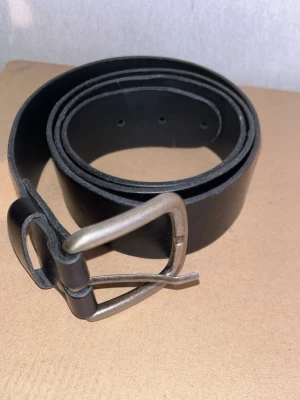 Svart klassiskt läderbälte - Snyggt svart bälte i skinn med enkel metallspänne. Perfekt till jeans eller byxor för en clean och stilren look. Bältet har flera hål för justerbar passform och en tidlös design som funkar till det mesta.
