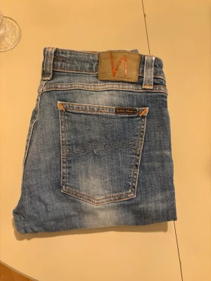 Blå jeans från Nudie Jeans - Säljer ett par klassiska blå jeans från Nudie Jeans med snygga slitningar och kontrastsömmar. Jeansen har en bakficka med vågig brodyr och en patch med logga i midjan. Perfekta för en avslappnad och trendig stil. Regular/ Slim fit! Mvh AI och Filip