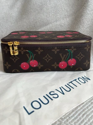 Louis Vuitton necessär med körsbär - Unik Louis Vuitton necessär i brunt monogrammönster med färgglada körsbärsdetaljer och guldigt blixtlås. Tillverkad i tåligt canvasmaterial och har en rektangulär form. Perfekt för att förvara smink eller småprylar. Mått 23x8x15