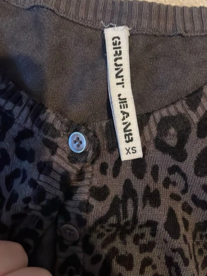 Leopardmönstrad kofta från Grunt Jeans - Snygg leopardmönstrad kofta från Grunt Jeans i svart och brun. Koftan har knappar framtill och ribbade kanter vid hals och ärmslut. Perfekt för dig som vill sticka ut med ett djurmönster i garderoben.