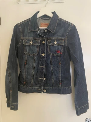 Mörkblå jeansjacka från Zoul - Snygg mörkblå jeansjacka från Zoul med klassisk krage, vita knappar och två bröstfickor med lock. Kontrastsömmar i orange och vit samt en liten röd detalj på ena fickan. Perfekt att slänga över en hoodie eller t-shirt. Använt väldigt lite - den är i väldigt bra skick! 
