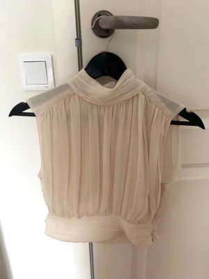 Beige plisserad ärmlös blus - Säljer en elegant beige ärmlös blus med plisserat tyg och hög krage. Blusen har en lätt transparent känsla och knäppning i nacken. Perfekt för dig som gillar en chic och stilren look. Den är i en Creamfärgad nyans