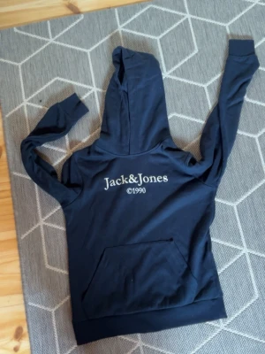 Mörkblå hoodie från Jack & Jones - Mörkblå hoodie från Jack & Jones med vit logga och årtal på ryggen. Klassisk huva och stor magficka framtill. Perfekt för en avslappnad och stilren look. Mjuk bomullskänsla och långa ärmar. strl 152 men passar allt från 152-160
