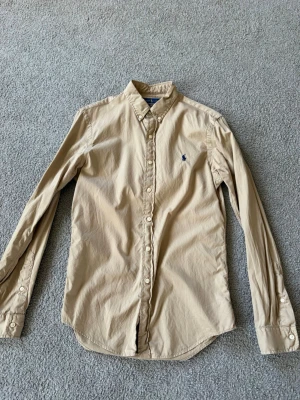 Beige skjorta från Ralph Lauren - Klassisk beige skjorta från Ralph Lauren med blå broderad logga på bröstet. Skjortan har lång ärm, knappar hela vägen och krage med knapp. Perfekt för en clean och stilren look.