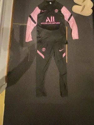 PSG träningsset svart/rosa Nike - Snyggt PSG träningsset från Nike i svart med rosa detaljer. Setet består av långärmad tröja och matchande byxor, båda med PSG-logga och Nike swoosh. Materialet är troligtvis polyester, perfekt för träning eller sport.