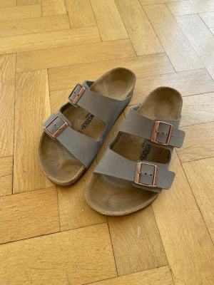 Grå Birkenstock sandaler med spännen - Stilrena grå sandaler från Birkenstock med två breda remmar och justerbara metallspännen i kopparfärg. Fotbädden är anatomiskt utformad och sulan är i slitstark gummi. Perfekta för en avslappnad och trendig look.