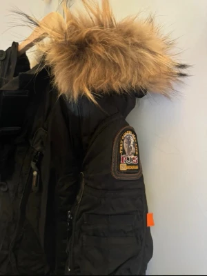 Svart Parajumpers parkas med pälskrage - Snygg svart parkas från Parajumpers med stor huva och fluffig pälskant. Jackan har flera fickor, dragkedja och knappar framtill samt klassisk Parajumpers-logga på ärmen. Perfekt för kalla dagar.
