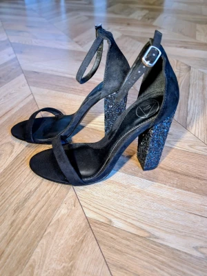 Svarta pumps med glitterklack - Stilrena svarta pumps med breda remmar över tårna och runt ankeln. Klackarna är höga och täckta av svart glitter som ger extra bling. Skorna har en justerbar rem med silverfärgat spänne. Materialet ser ut att vara mockaimitation.