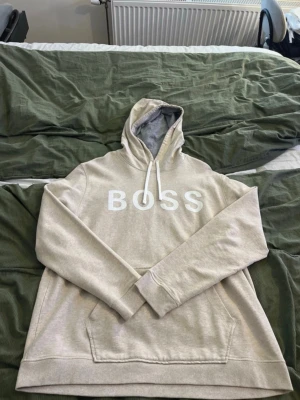 Beige hoodie från BOSS med tryck - Snygg beige hoodie från BOSS med stort vitt logotryck på bröstet. Hoodien har huva med vita dragsnören och en klassisk känguruficka framtill. Mjuk insida och ribbade muddar vid ärmslut och nederkant. Perfekt för en chill och stilren look.