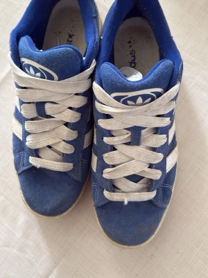 Blå Adidas Campus sneakers - Säljer ett par klassiska Adidas Campus sneakers i blå mocka med vita läderdetaljer och de ikoniska tre ränderna på sidan. Skorna har vita snören och en låg silhuett. Perfekta för dig som gillar retrostil och streetwear.