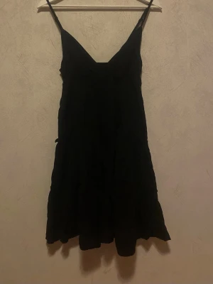 Svart kort klänning med smala axelband - Svart kort klänning i storlek S med smala axelband och v-ringning. Klänningen har en lätt volang nertill och är tillverkad i ett tunt, mjukt tyg som ger en luftig känsla. Perfekt för varma dagar eller när du vill ha en enkel och stilren look.