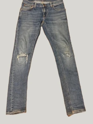 Blå nudie jeans med snygga slitningar  - Säljer ett par blå jeans med passformen slim fit och slitningar på båda knäna. Jeansen har klassisk femficksdesign och ljus tvätt med snygga detaljer. Perfekta för en avslappnad och trendig look. MODELL: skinny lin 