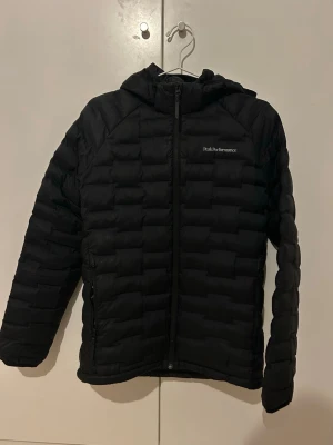 Svart pufferjacka från Peak Performance( lättviktsdunjacka) stl 170) - Snygg svart pufferjacka från Peak Performance med huva och dragkedja framtill. Jackan har quiltad design och diskret logga på bröstet. Perfekt för kalla dagar och enkel att matcha med olika outfits.