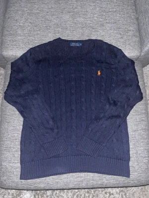 Mörkblå kabelstickad tröja Polo Ralph Lauren - Snygg mörkblå kabelstickad tröja från Polo Ralph Lauren med klassisk orange logga på bröstet. Tröjan har rund hals, ribbade muddar och är gjord i mjukt stickat material. Perfekt för dig som gillar stilrena och tidlösa plagg.