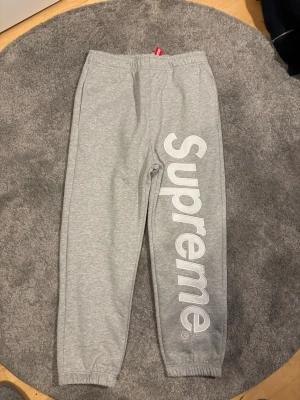 Gråa Supreme mjukisbyxor - Säljer ett par gråa mjukisbyxor från Supreme med stort vitt logotryck längs ena benet. Byxorna har resår i midjan och vid bensluten samt en bakficka. Perfekta för en chill och streetwear-inspirerad look.