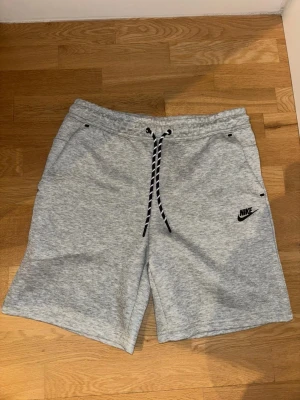 Grå sweatshorts från Nike med snörning - Grå mjuka sweatshorts från Nike med svart logga på vänster ben. Shortsen har snörning i midjan och två sidofickor. Tillverkade i bomullsmix för en bekväm känsla. Perfekta för chill eller träning.